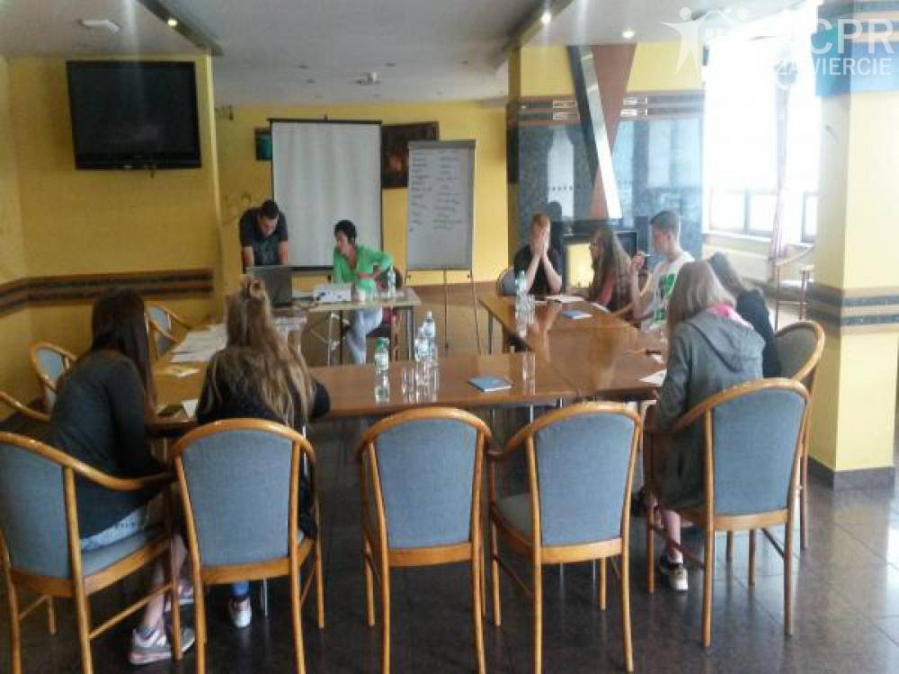 Zdjęcie: Uczestniczki, uczestnicy i prowadząca siedzą wokół stołu w sali warsztatowej